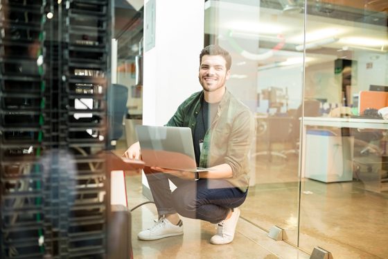 portrait-happy-young-male-network-engineer-with-laptop-hand-working-datacenter